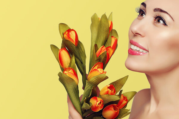 Spring beauty model studio shooting. Close-up portrait of smiling young beautiful woman with flowers orange tulips on yellow background. Fashion fresh natural makeup. Romantic style. Tenderness.