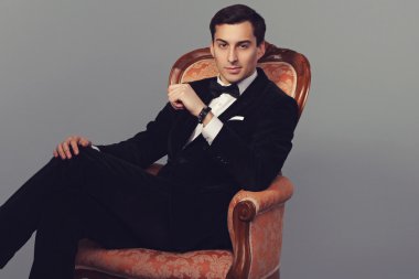 Kendine güvenen başarılı genç yakışıklı adam işadamı vintage gri arka koltukta oturan papyon ile şık elbiseli. Erkeklik. Erkek güzellik. Moda modeli studio vurdu. İtalyan tarzı. Lüks.