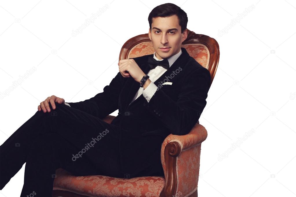 Confident successful young handsome man businessman in elegant suit with bow tie sitting on vintage armchair on white background. Manhood. Male beauty. Fashion model studio shot. Italian style.