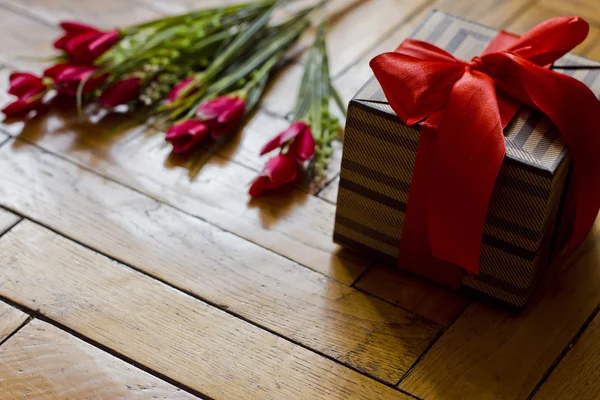 Открытая подарочная коробка с красным бантом и бежевыми коричневыми полосками на деревянном фоне. Букет красных алых цветов тюльпаны. Романтический стиль. Праздники . — стоковое фото