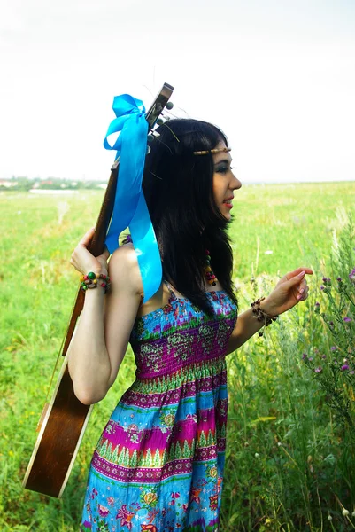 Hermosa joven de hippie en vestido azul violeta con la guitarra azul raya en la hierba mirando a la distancia. Disfruta de la naturaleza. Estilo bohemio. Boho chic. Libertad. Paz. . — Foto de Stock