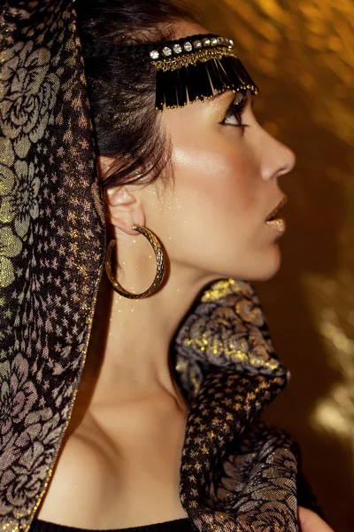 Şal hijab etnik aksesuarları küpe ve kolye ile esmer genç güzel Arap kadın. Altın arka plan üzerinde kız profil. Çekim modeli. Altın moda makyaj. Lüks. Zerafet. Duygusallık. Oryantal tarzda.