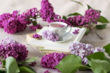 Romantik arka plan, bir fincan çay, leylak çiçekleri ve beyaz masanın üzerinde açık bir kitap.