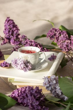 Romantik arka plan, bir fincan çay, leylak çiçekleri ve beyaz masanın üzerinde açık bir kitap.