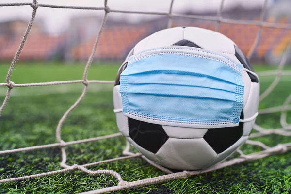 Futbol Gol Ağı yakınlarında Siyah ve Beyaz Futbol Topu Tıbbi Maske 'de. Arka planda Stadyumun Boş Koltukları var. Futbol müsabakaları durduruldu. COVID-19 Konsepti