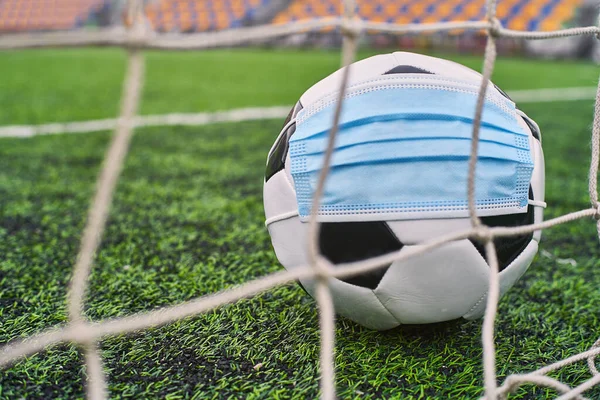 Futbol Gol Ağı yakınlarında Siyah ve Beyaz Futbol Topu Tıbbi Maske 'de. Arka planda Stadyumun Boş Koltukları var. Futbol müsabakaları durduruldu. COVID-19 Konsepti
