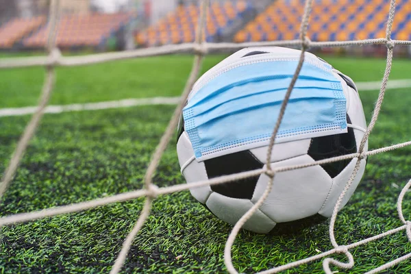 Futbol Gol Ağı yakınlarında Siyah ve Beyaz Futbol Topu Tıbbi Maske 'de. Arka planda Stadyumun Boş Koltukları var. Futbol müsabakaları durduruldu. COVID-19 Konsepti