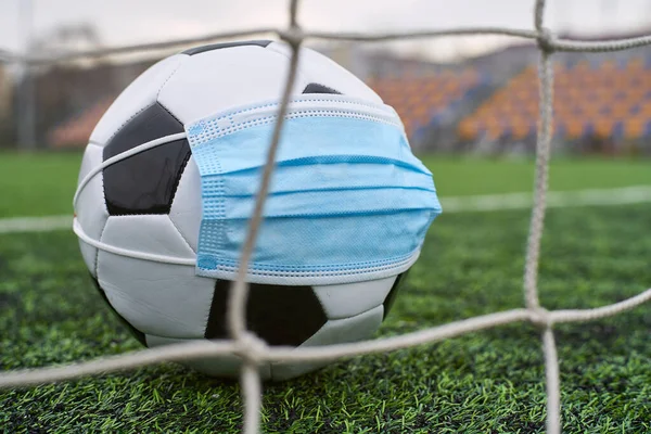 Futbol Gol Ağı yakınlarında Siyah ve Beyaz Futbol Topu Tıbbi Maske 'de. Arka planda Stadyumun Boş Koltukları var. Futbol müsabakaları durduruldu. COVID-19 Konsepti