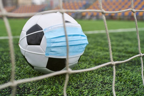 Futbol Gol Ağı yakınlarında Siyah ve Beyaz Futbol Topu Tıbbi Maske 'de. Arka planda Stadyumun Boş Koltukları var. Futbol müsabakaları durduruldu. COVID-19 Konsepti