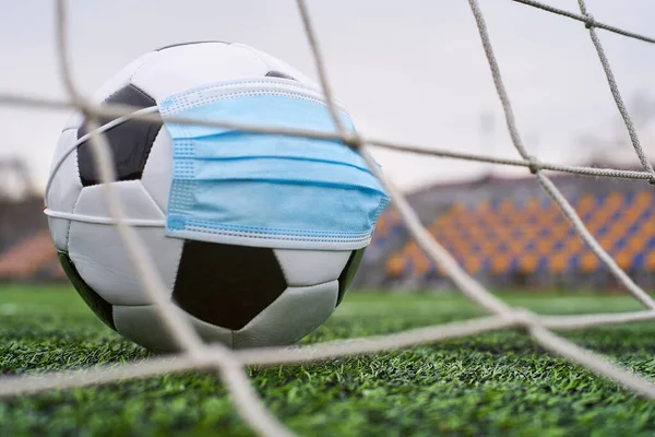 Futbol Gol Ağı yakınlarında Siyah ve Beyaz Futbol Topu Tıbbi Maske 'de. Arka planda Stadyumun Boş Koltukları var. Futbol müsabakaları durduruldu. COVID-19 Konsepti