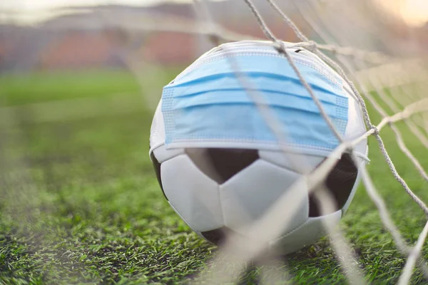 Soccer Ball in Medical Mask on Stadium Green Grass. Ball in Goal Net. Football Competitions Stopped