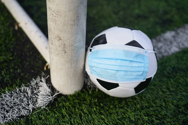 Black and White Soccer Ball in Medical Mask Lying Near Soccer Goal Post. Empty Stadium. Football Competitions During Quarantine Time. COVID-19 Concept