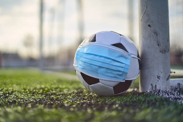 Black and White Soccer Ball in Medical Mask Lying Near Soccer Goal Post. Empty Stadium. Football Competitions During Quarantine Time. COVID-19 Concept