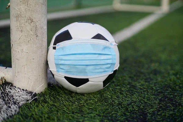 Black and White Soccer Ball in Medical Mask Lying Near Soccer Goal Post. Empty Stadium. Football Competitions During Quarantine Time. COVID-19 Concept