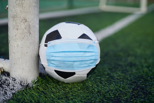 Black and White Soccer Ball in Medical Mask Lying Near Soccer Goal Post. Empty Stadium. Football Competitions During Quarantine Time. COVID-19 Concept