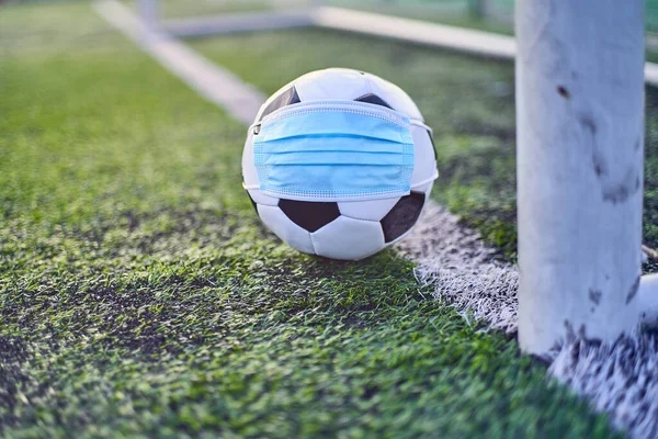 Black and White Soccer Ball in Medical Mask Lying Near Soccer Goal Post. Empty Stadium. Football Competitions During Quarantine Time. COVID-19 Concept