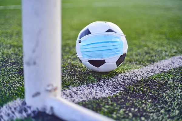 Black and White Soccer Ball in Medical Mask Lying Near Soccer Goal Post. Empty Stadium. Football Competitions During Quarantine Time. COVID-19 Concept