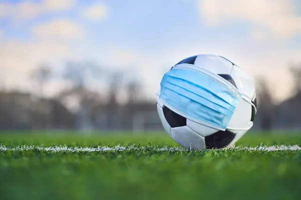 Medikal Maskeli Siyah ve Beyaz Futbol Balosu Stadyumun Çimlerinde Yatar. Arka plan Boş Stadyum ve Gökyüzü. Karantina zamanında futbol müsabakaları. COVID-19 Konsepti