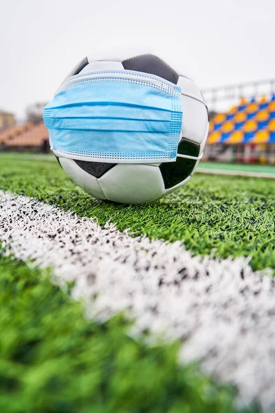Boş stadyumda futbol topu. Koronavirüsten korunmak için tıbbi maskeli futbol topu, insanlar olmadan plastik koltuklardan korunmak için. Dikey fotoğraf