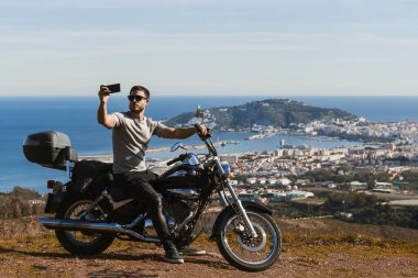 Motosikletin üzerinde oturan bisikletçi, cep telefonuyla manzarayla selfie çekiyor.