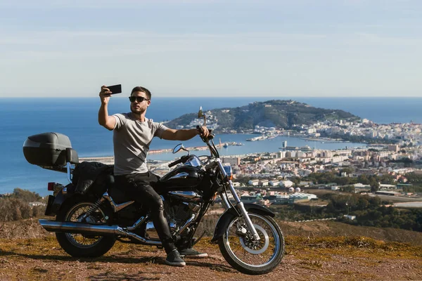 Motosikletin üzerinde oturan bisikletçi, cep telefonuyla manzarayla selfie çekiyor.