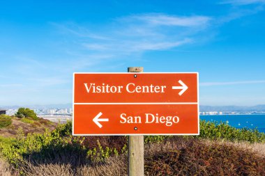 San Diego ve Ziyaretçi Merkezi kahverengi yol işareti beyaz harfli ve yönlü oklarla. Arka plan San Diego Körfezi ve Şehir