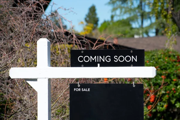 Coming Soon sign near a house for sale in a residential neighborhood. Green trees background.