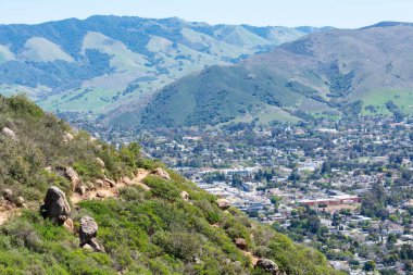 Aerial view of Bishop Peak trail surrounded by chaparral on the hill. Background San Luis Obispo area sprawl and scenic green mountains of Santa Lucia Range clipart