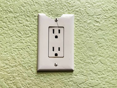 Broken electrical socket with chipped cover plate and textured green stucco wall background, showing need for replacement and illustrating safety inspection or DIY home repair