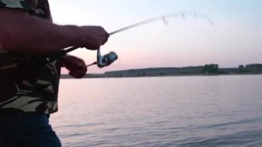 A fisherman turns the handle of a spinning reel at sunset. A person who is fond of fishing on the lake, tightens the fishing line with a reel of fish in the summer.