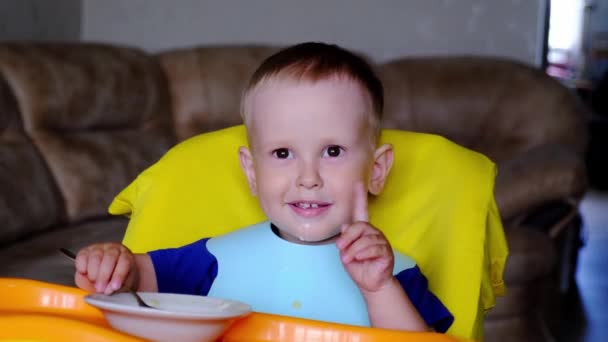 2歳の男の子は 子供用の座席からスプーンでミルクスープを食べるのを楽しんでいます 子供は自分で皿から食べることを学ぶ — ストック動画