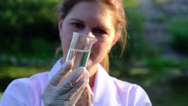 Egy laboratóriumi asszisztens lány tanulmányozza a vízmintákat egy lombikban, a tó partján, az ökológia fogalmát. — Stock videók