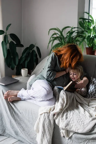 Happy pregnant mom reads a book to her older daughter. at home on the couch among houseplants. Happy family, second child, planning, activities with the child, development, reading a book, together.
