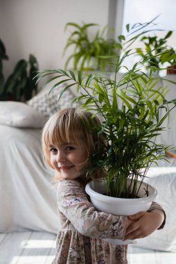 Ev bitkileriyle ilgilenen küçük bir kız çocuğu. Çocuk gelişimi, hobiler, yaşam tarzı, bitkilere duyulan sevgi, çocuğun uyumlu gelişimi. Evde bahçıvanlık, turuncu önlüklü küçük bahçıvan..