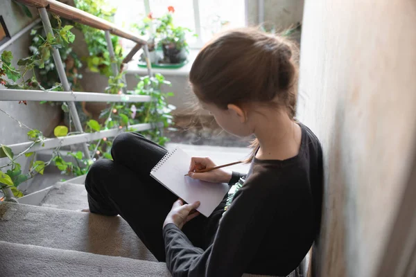 A teenage girl sits on the stairs of the house and draws with a pencil in a notebook. Children and creativity, home education during quarantine, child development through creativity.