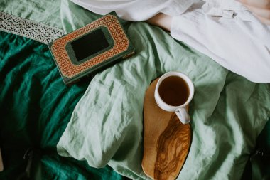 Kadın eli sıcak bir fincan çayı yatakta tutar, çarşaf örtüsü, köy hayatı, yatakta okumak, rahatlama, sükunet, dijital detoks.