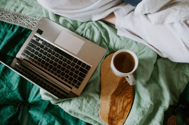 Yatak örtüleri ve ahşap tepside bir fincan sıcak çay olan bir dizüstü bilgisayar. Bilinçli yaşam konsepti, evden çalışma, uzaktan çalışma, serbest çalışma, köy hayatı.