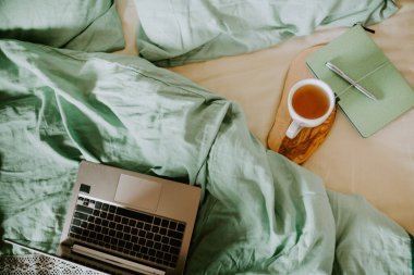 Yatak örtüleri ve ahşap tepside bir fincan sıcak çay olan bir dizüstü bilgisayar. Bilinçli yaşam konsepti, evden çalışma, uzaktan çalışma, serbest çalışma, köy hayatı.