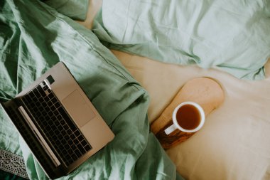 Yatak örtüleri ve ahşap tepside bir fincan sıcak çay olan bir dizüstü bilgisayar. Bilinçli yaşam konsepti, evden çalışma, uzaktan çalışma, serbest çalışma, köy hayatı.