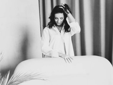 Siyah saçlı, beyaz gömlekli çekici bir kadının portresi. Evdeki kanepenin yanında duruyor. Sabah konsepti. Stüdyo çekimi