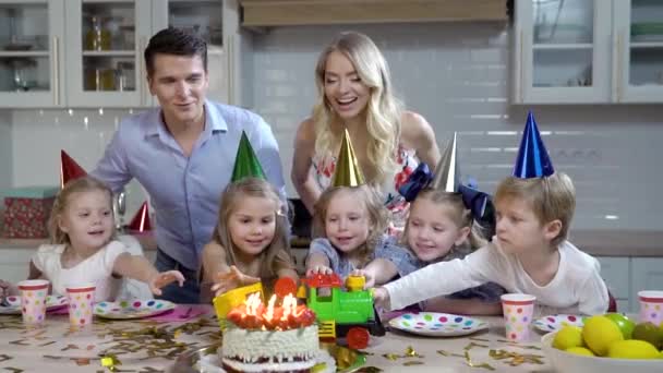 Szczęśliwa rodzina pięciorga dzieci, tata i mama świętują urodziny córek w kuchni. Rodzice dają jej zabawkowy pociąg. Wszystkie ubrane w świąteczne ubrania, na głowach czapki dla dzieci. — Wideo stockowe
