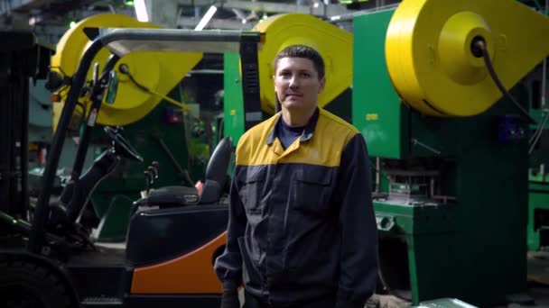 Porträt eines jungen Mitarbeiters in Uniform des Unternehmens, der Fabrik, des Lagers auf dem Hintergrund des Laders. Er blickt in die Kamera und lächelt — Stockvideo