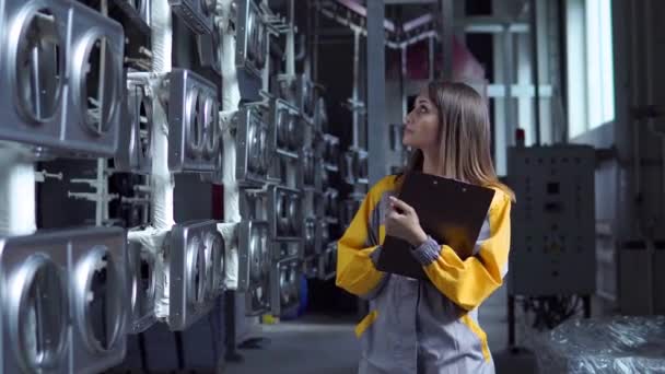 En ung söt vit kvinna arbetar i fabrikens målarverkstad, kontrollerar de omålade metallprodukterna och gör rekord i checklistan. — Stockvideo