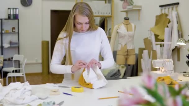 Mujer con el pelo rubio freelancer, diseñador de moda o sastre que trabaja en un diseño o proyecto con telas de colores en el taller. Vista frontal — Vídeo de stock