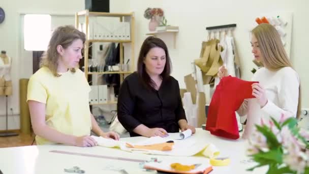 Tre kvinner i verkstedet på klesfabrikken. De ser på fargede stoffer for en ny kjole og ser etter vellykkede kombinasjoner av tekstiler. De smiler og har det gøy – stockvideo