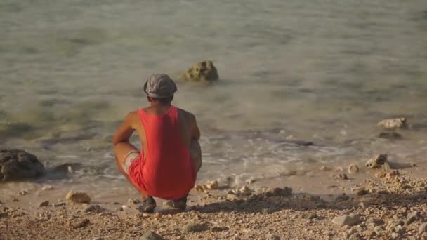 Bakifrån av en ung svart man, sitter på hans lår på stranden och touch sand och vatten. — Stockvideo