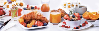 İçinde kruvasan, haşlanmış yumurta, böğürtlenli granola, taze portakal suyu, kahve ve beyaz bir masada papatya bulunan zengin bir kahvaltı panoraması..