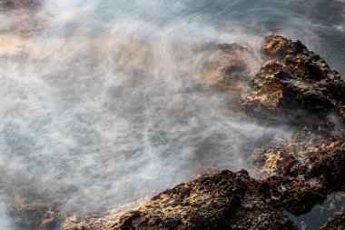 Gün batımında deniz suyu, hareket halinde bulanıklık, sahilde taşlar. Atlantik Okyanusu, Tenerife Adası. Uzun pozlama.