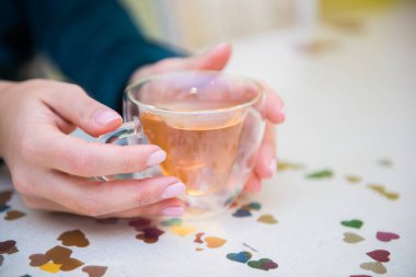 Masanın yanında oturan şeffaf bir fincanı yeşil çayla tutan bir kızın yan görüntüsü. Bardaktaki parlak röfleler. Samimi bir resim. Kapatın. Organik sağlık hizmetleri.