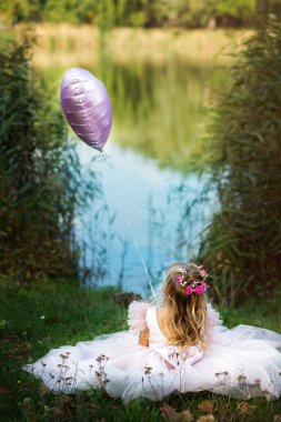 Güzel, yüzü olmayan, pembe elbiseli sarışın bir kızın dikey resmi. Orman ve göl manzarasına karşı doğada oturuyor. Çocuk elinde balon tutuyor. Umut ve mutluluk.